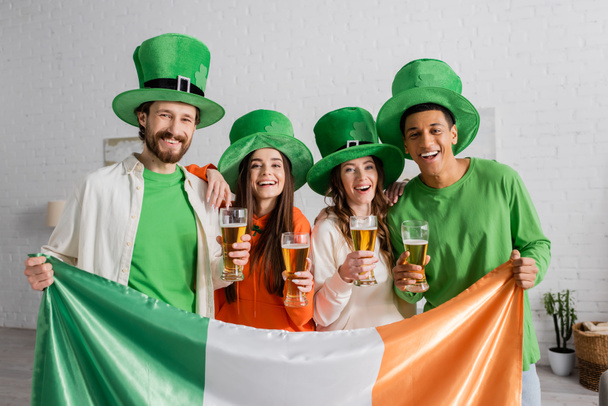 radosnych i wielokulturowych przyjaciół w zielonych kapeluszach trzymających kieliszki piwa i irlandzką flagę podczas obchodów Święta Patryka  - Zdjęcie, obraz