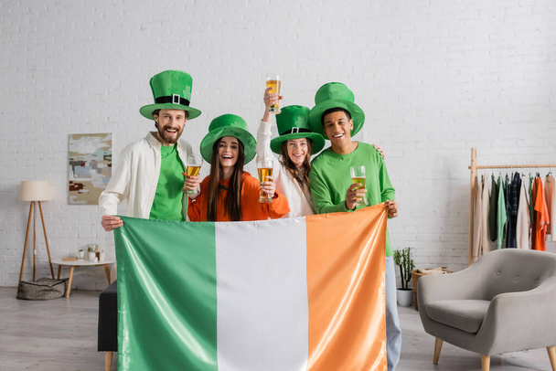 boldog és multikulturális barátok zöld kalapban, akik egy pohár sört és ír zászlót tartanak, miközben Szent Patrik napot ünneplik  - Fotó, kép