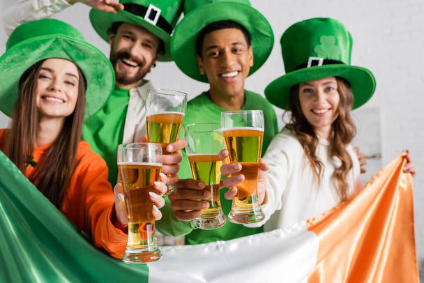 веселые и мультикультурные друзья в зеленых шляпах, держащие бокалы пива и ирландский флаг во время празднования Дня Святого Патрика  - Фото, изображение