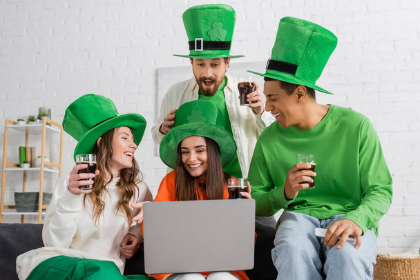 позитивні та багатоетнічні друзі в зелених капелюхах тримають келихи темного пива, дивлячись на ноутбук
  - Фото, зображення