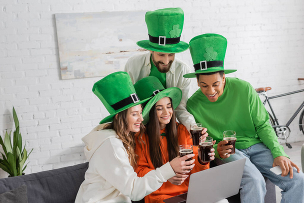 happy and multiethnic friends in green hats clinking glasses of dark beer while looking at laptop  - 写真・画像