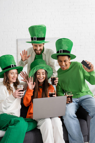 amis heureux et multiethniques dans des chapeaux verts tenant des verres de bière sombre tout en agitant les mains à l'ordinateur portable pendant l'appel vidéo  - Photo, image