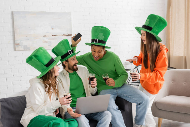 amigos alegres y multiétnicos en sombreros verdes sosteniendo vasos de cerveza oscura durante la celebración del Día de San Patricio en casa - Foto, imagen