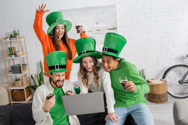 cheerful woman holding laptop near multiethnic friends with glasses of dark beer during video call on Saint Patrick Day - Zdjęcie, obraz