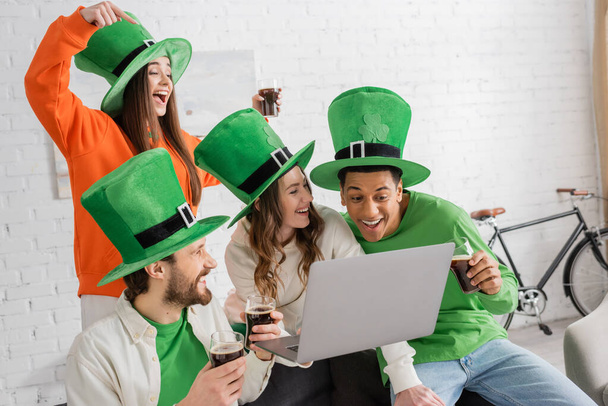 cheerful woman holding laptop near multiethnic friends with glasses of dark beer on Saint Patrick Day - Zdjęcie, obraz