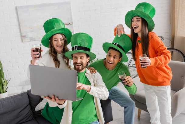 bearded man holding laptop near joyful multiethnic friends with glasses of dark beer while celebrating Saint Patrick Day  - 写真・画像