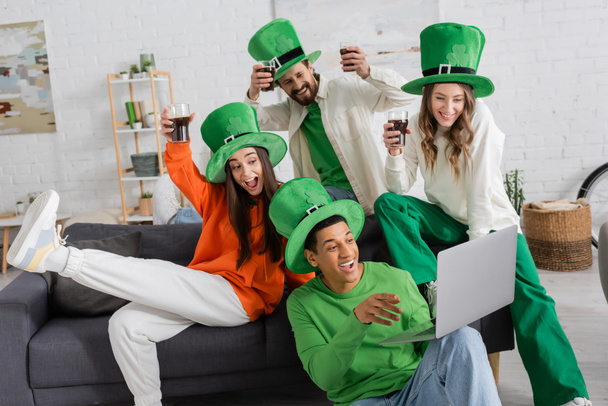 happy african american man holding laptop during video call near cheerful friends toasting glasses of beer on Saint Patrick Day - Valokuva, kuva