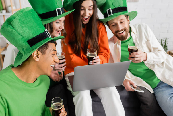 podekscytowani przyjaciele międzyrasowe patrząc na laptopa podczas rozmowy wideo trzymając szklanki ciemnego piwa na Saint Patrick Day - Zdjęcie, obraz