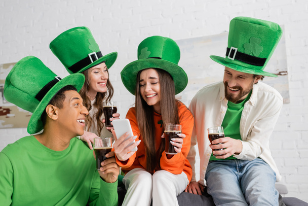 счастливые межрасовые друзья смотрят на друга со смартфона, держа в руках стаканы тёмного пива в День Святого Патрика - Фото, изображение