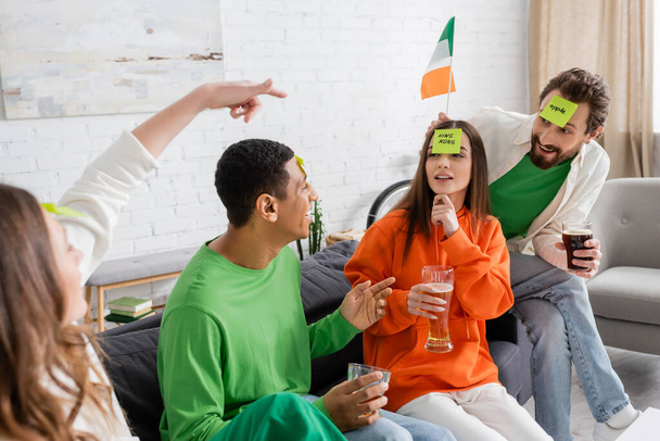 woman pointing at friends with sticky notes on foreheads near Irish flag while playing guess who game on Saint Patrick Day - Photo, Image