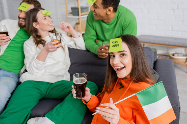 схвильована жінка з королем на липкій ноті з ірландським прапором і склянкою пива біля міжрасових друзів у День святого Патрика. - Фото, зображення