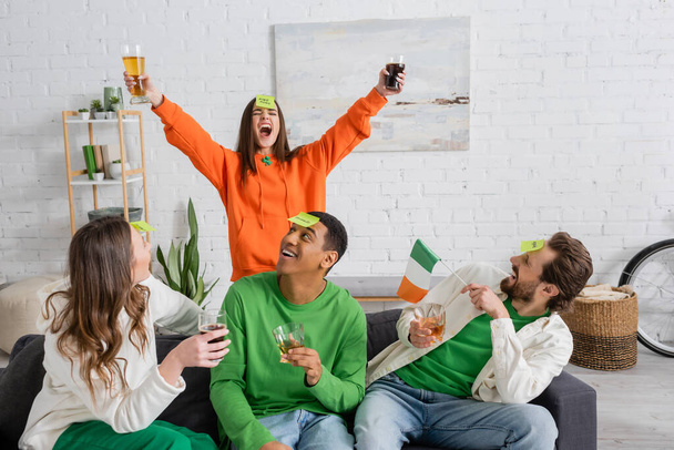 Excited woman holding beer near interracial friends with irish flag playing who i am at home  - Photo, image