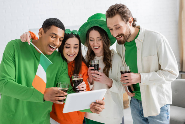 Позитивные многоэтнические друзья с пивом, празднуя День святого Патрика и используя цифровые планшеты дома  - Фото, изображение
