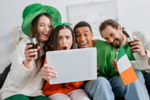 Размытые межрасовые друзья используют цифровые планшеты, празднуя День святого Патрика дома  - Фото, изображение