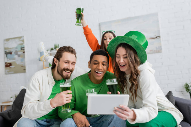 Excited woman holding green beer and digital tablet near multiethnic friends at home  - 写真・画像