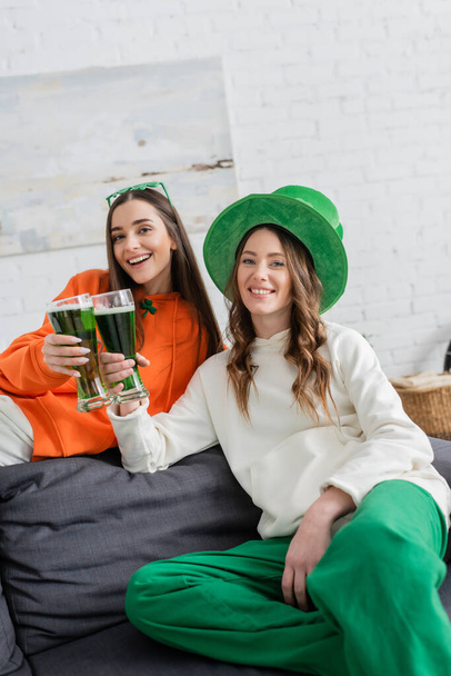 Χαμογελώντας νεαρές γυναίκες κρατώντας πράσινη μπύρα και κοιτάζοντας την κάμερα στο σπίτι  - Φωτογραφία, εικόνα