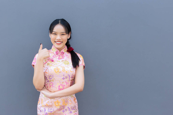 Beautiful Asian woman with long hair who wears pink Cheongsam dress in Chinese new year theme while her hand shows to present something and showing hand as thump up and looks at camera smiling happily with the grey background. - Photo, Image