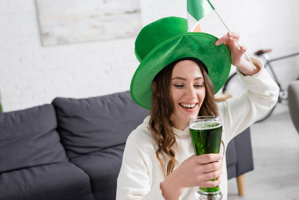 Fröhliche Frau mit Hut und grünem Bier und irischer Flagge zu Hause  - Foto, Bild