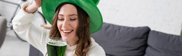 Θετική νεαρή γυναίκα στο καπέλο κρατώντας ποτήρι πράσινη μπύρα στο σπίτι, πανό  - Φωτογραφία, εικόνα
