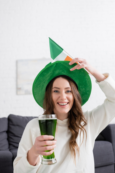Mujer positiva con sombrero sosteniendo cerveza verde y bandera irlandesa mientras mira la cámara en casa  - Foto, imagen