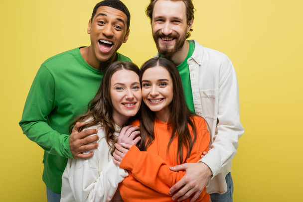 Cheerful interracial men hugging friends isolated on yellow  - Photo, Image
