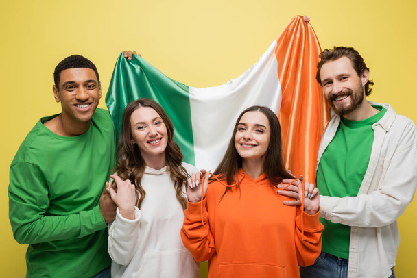Sonrientes hombres multiétnicos sosteniendo bandera irlandesa cerca de amigos aislados en amarillo  - Foto, imagen