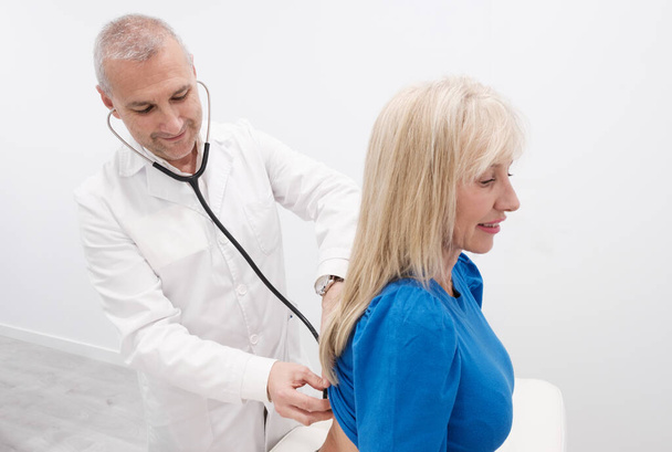Ένας έμπειρος γιατρός κάνει ένα check-up σε μια ώριμη γυναίκα ασθενή χρησιμοποιώντας ένα στηθοσκόπιο σε ένα σύγχρονο, καθαρό, κλινικό περιβάλλον με λευκό φόντο. Έννοια: υγειονομική περίθαλψη και σύστημα δημόσιας υγείας - Φωτογραφία, εικόνα