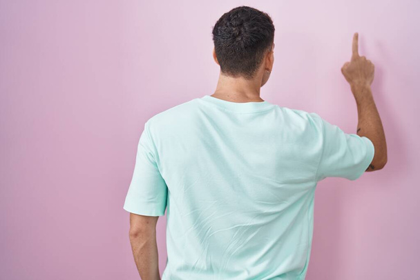 Knappe Spaanse man die over roze achtergrond staat en achteruit wijst met de vinger in de hand  - Foto, afbeelding