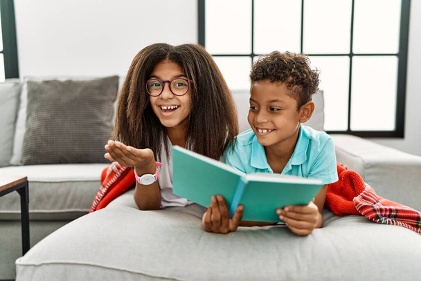 Dwoje rodzeństwa leżących na kanapie czytających książkę skierowaną na bok z otwartymi dłońmi pokazującymi przestrzeń do kopiowania, prezentujących uśmiechniętą reklamę podekscytowaną radością  - Zdjęcie, obraz