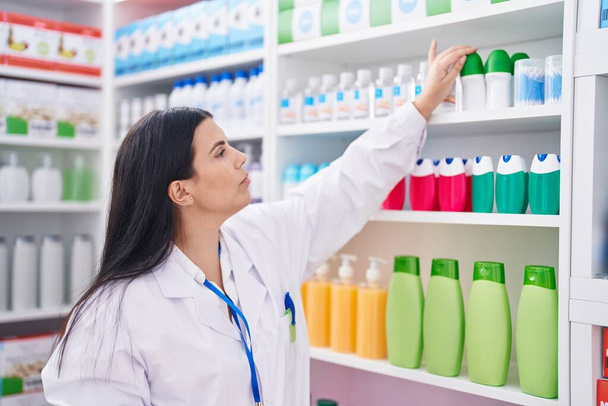 Joven hermosa mujer hispana farmacéutica sosteniendo el producto en estanterías en la farmacia - Foto, Imagen