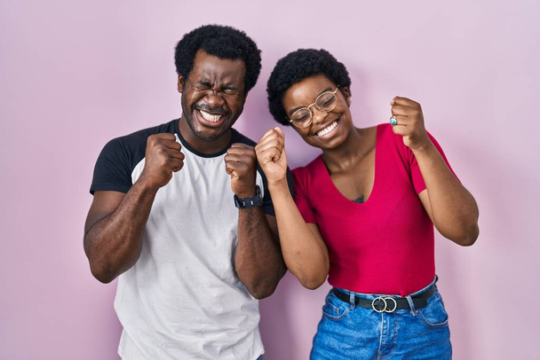Молода афроамериканська пара, що стоїть над рожевим фоном, захоплюється успіхом з піднятими руками і закриває очі, що святкують перемогу, посміхаючись. поняття переможця.  - Фото, зображення