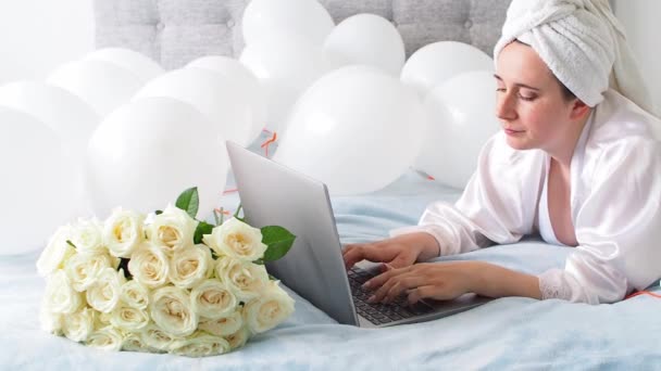 Woman in white clothes and towel on her head lying on a bed with white color balloons and bouquet of roses. Woman works at home, using her laptop, computer. Birthday, wedding. Copy space. Preparation. - Filmati, video