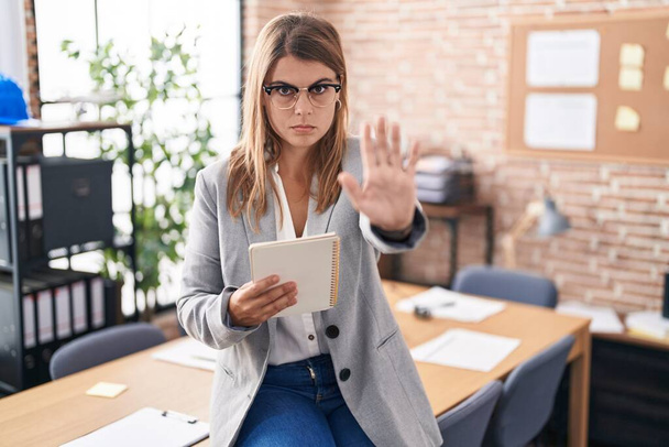 De jonge Spaanse vrouw die op kantoor werkt en een bril draagt, zingt niet meer met een handpalm. waarschuwingsuitdrukking met negatief en ernstig gebaar op het gezicht.  - Foto, afbeelding