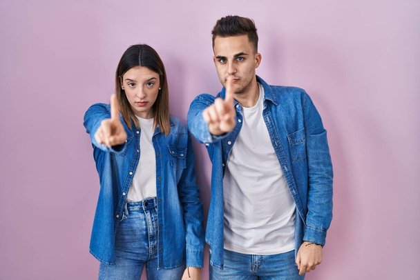Giovane coppia ispanica in piedi su sfondo rosa che punta con il dito verso l'alto e l'espressione arrabbiata, mostrando nessun gesto  - Foto, immagini