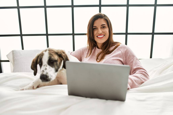Νεαρή γυναίκα που χρησιμοποιεί φορητό υπολογιστή αγκαλιάζει το σκυλί κάθεται στο κρεβάτι στο υπνοδωμάτιο - Φωτογραφία, εικόνα