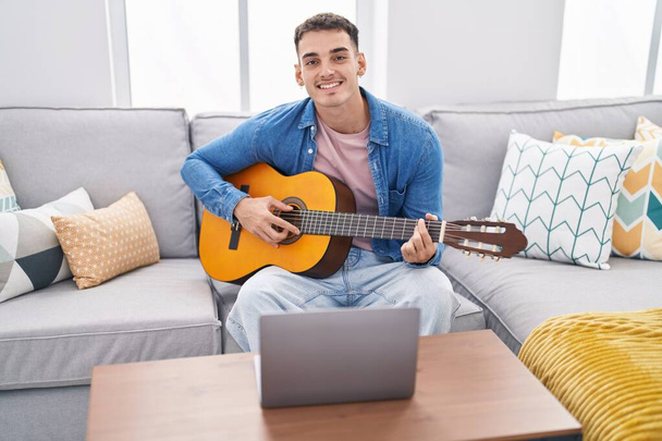 Hombre hispano joven que tiene clase de guitarra clásica en línea sentado en el sofá en casa - Foto, Imagen