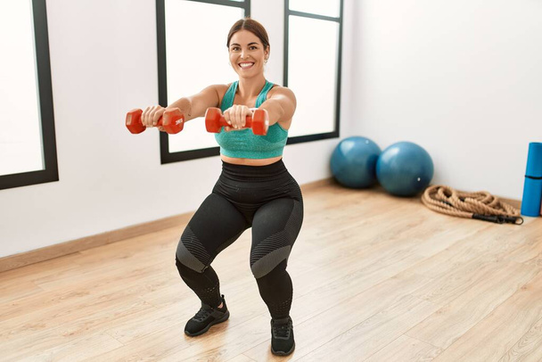 Fiatal gyönyörű spanyol nő mosolyog magabiztos segítségével súlyzók edzés a sport központ - Fotó, kép