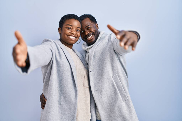 青い背景の上に立っている若いアフリカ系アメリカ人のカップルは、抱擁のために開いて腕で笑顔カメラを見ています。幸せを受け入れる陽気な表情.  - 写真・画像