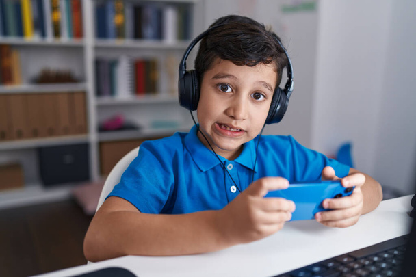 Adorable hispanic boy student using smartphone and headphones at classroom - 写真・画像