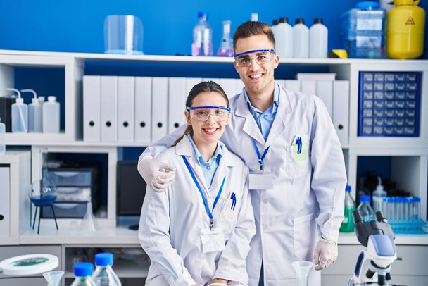 Мужчина и женщина ученые улыбаются уверенно обнимая друг друга в лаборатории - Фото, изображение