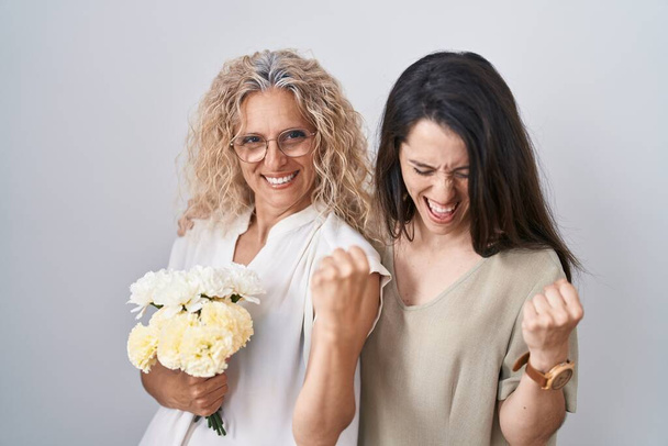 母親と娘の白い花の花束を保持非常に満足し、腕を上げて勝者のジェスチャーを行う興奮し、笑顔と成功のために叫んでいる。お祝いのコンセプト.  - 写真・画像