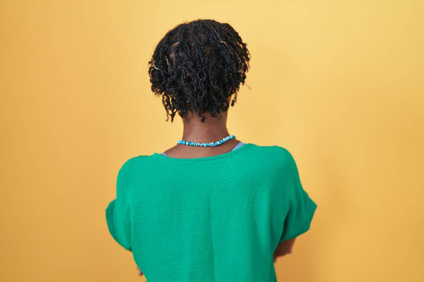 Afrikkalainen nainen rastat seisoo yli keltainen tausta seisoo taaksepäin katsoen pois ristissä kädet  - Valokuva, kuva