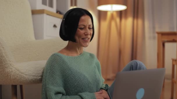 Happy friendly middle-aged woman in wireless headphones talking on video call on laptop, waving hello to webcam, talking, smiling, listening, nodding, sitting on floor at home. High quality 4k footage - Séquence, vidéo
