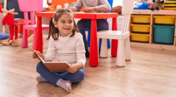 Чарівна іспаномовна дівчина читає книгу, сидячи на підлозі в дитячому садку
 - Фото, зображення