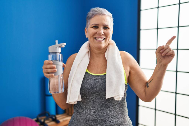 Жінка середнього віку в спортивному одязі та рушнику тримає воду усміхненою щасливою, вказуючи рукою та пальцем на бік
  - Фото, зображення