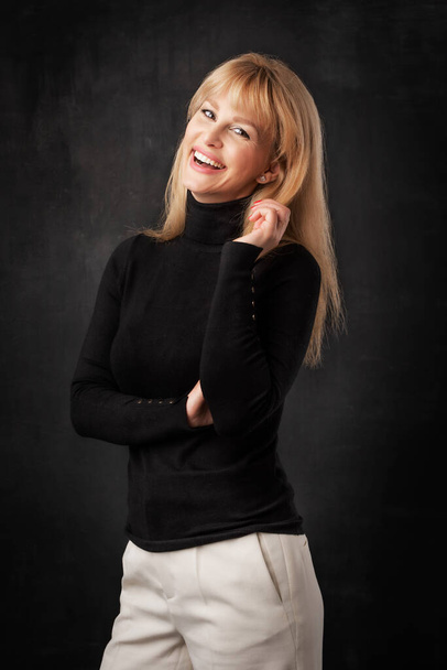 Beautiful smiling woman standing at isolated dark background. Blond haired woman wearing turtleneck sweater. Copy space.  - Foto, Imagem