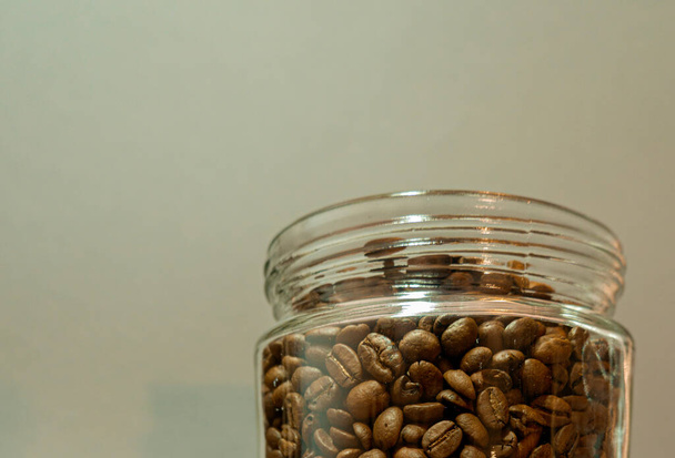 Un recipiente de vidrio lleno de granos de café tostados sobre un fondo gris cálido  - Foto, Imagen
