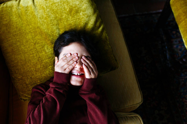 górny widok na ciemnowłose dziecko okrywające oczy na żółtej sofie - Zdjęcie, obraz