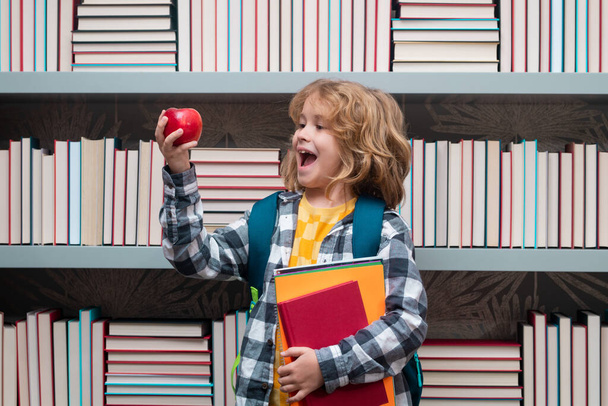 Iskolás fiú könyvekkel és almával a könyvtárban. Iskolás gyerek tanul az általános iskolában. Egy általános iskolai órán tanuló gyerek. - Fotó, kép