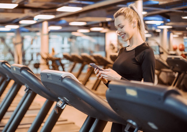 Fitness model listening music on smart phone charching positive energy before workout. Healthy fitness lifestyle concept with copy space. Athletic Sports Woman in Gym Puts on Wireless Headphones. - 写真・画像